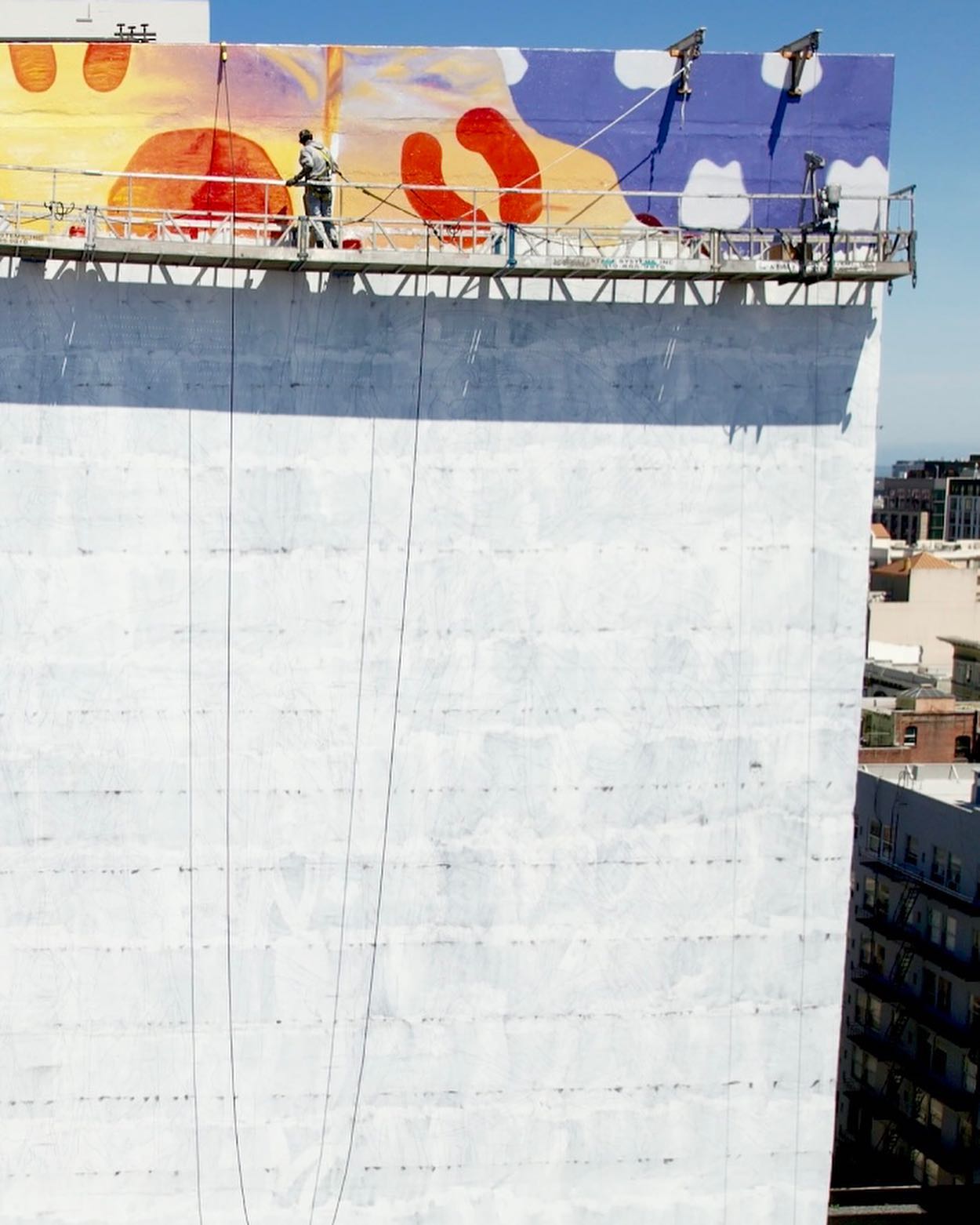 Hoist Media artist on suspended scaffold painting top section of large-scale mural on San Francisco building
