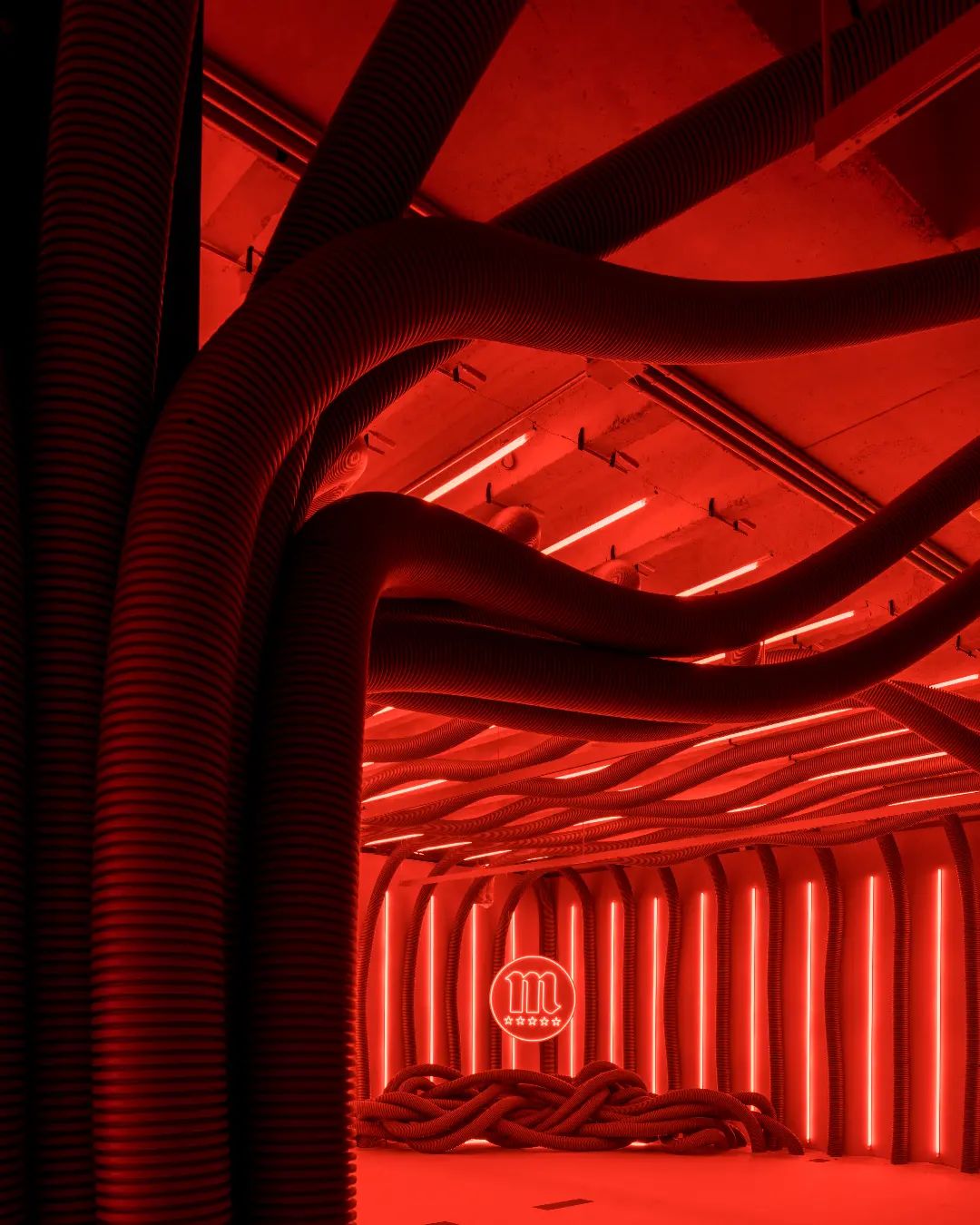 Dark angle view down red hose tunnel toward illuminated Mahou neon sign in Mahou Beer spatial design Madrid